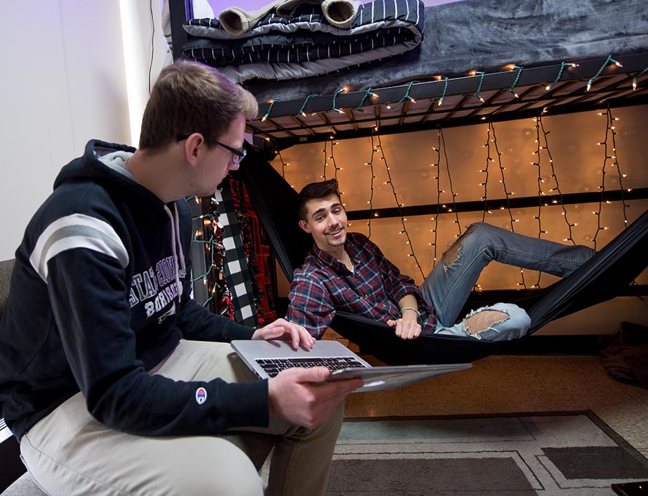 两个男孩在宿舍里, 一个睡在阁楼床下的吊床上，另一个坐在办公椅上，手里拿着一台笔记本电脑.